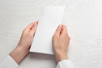 Young woman holding blank brochure at white wooden table, closeup. Mock up for design