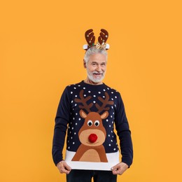 Photo of Senior man in reindeer headband showing his Christmas sweater on orange background