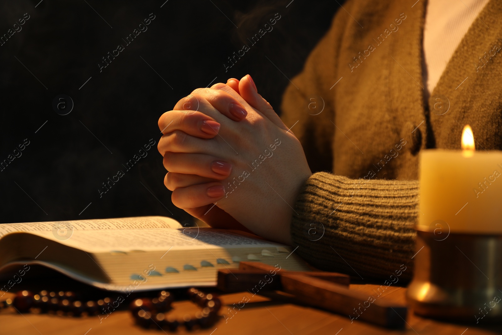 Photo of Woman praying at table with burning candle and Bible, closeup