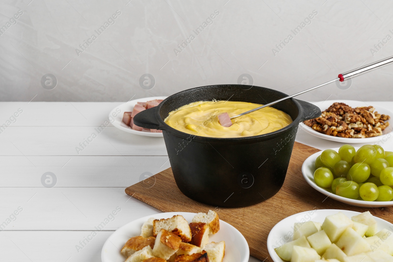 Photo of Fondue pot with tasty melted cheese, fork and different snacks on white wooden table, space for text