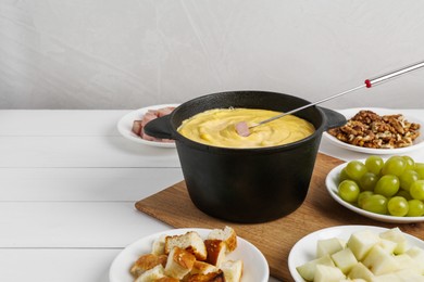 Fondue pot with tasty melted cheese, fork and different snacks on white wooden table, space for text