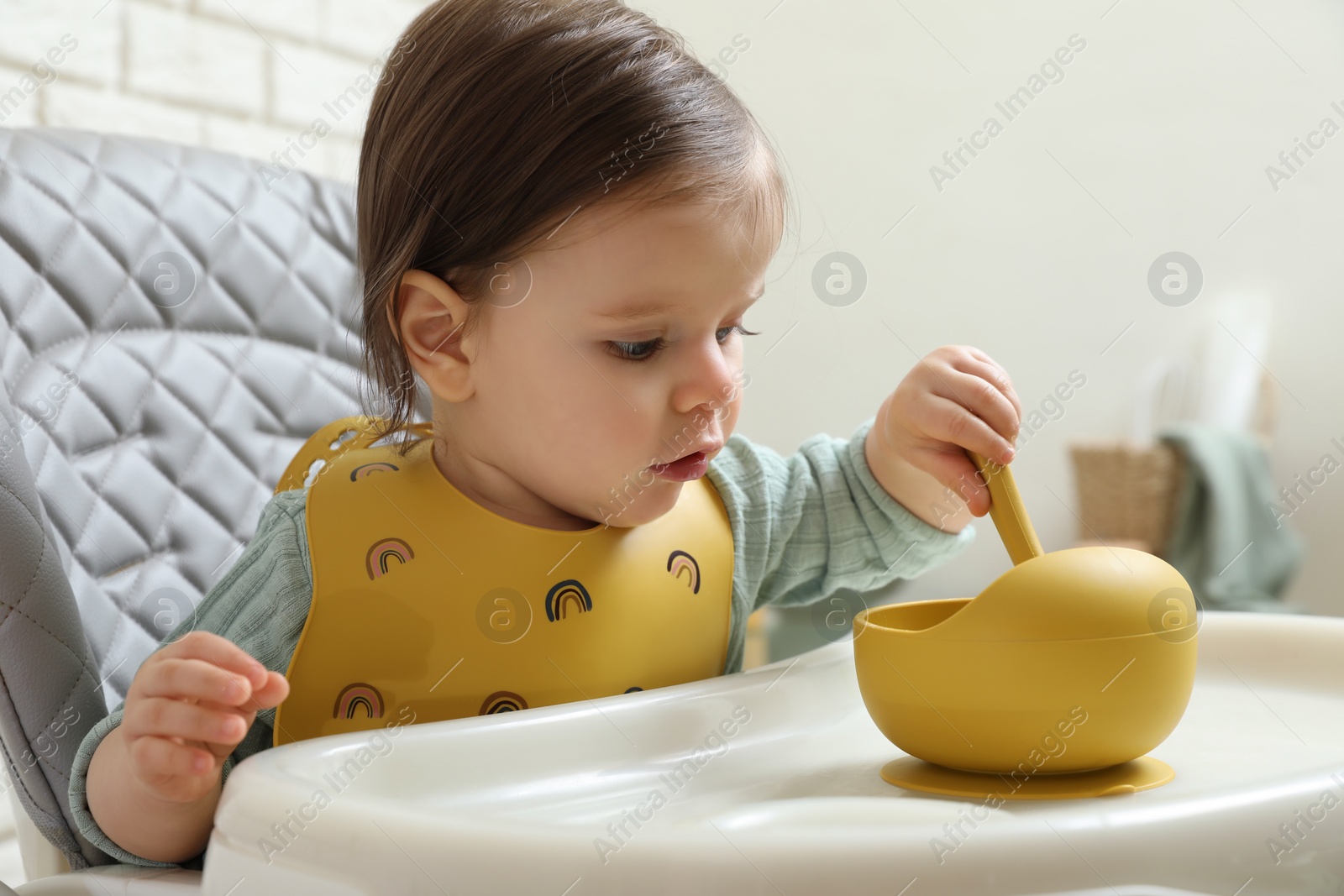 Photo of Cute little baby with spoon and bowl sitting in high chair indoors