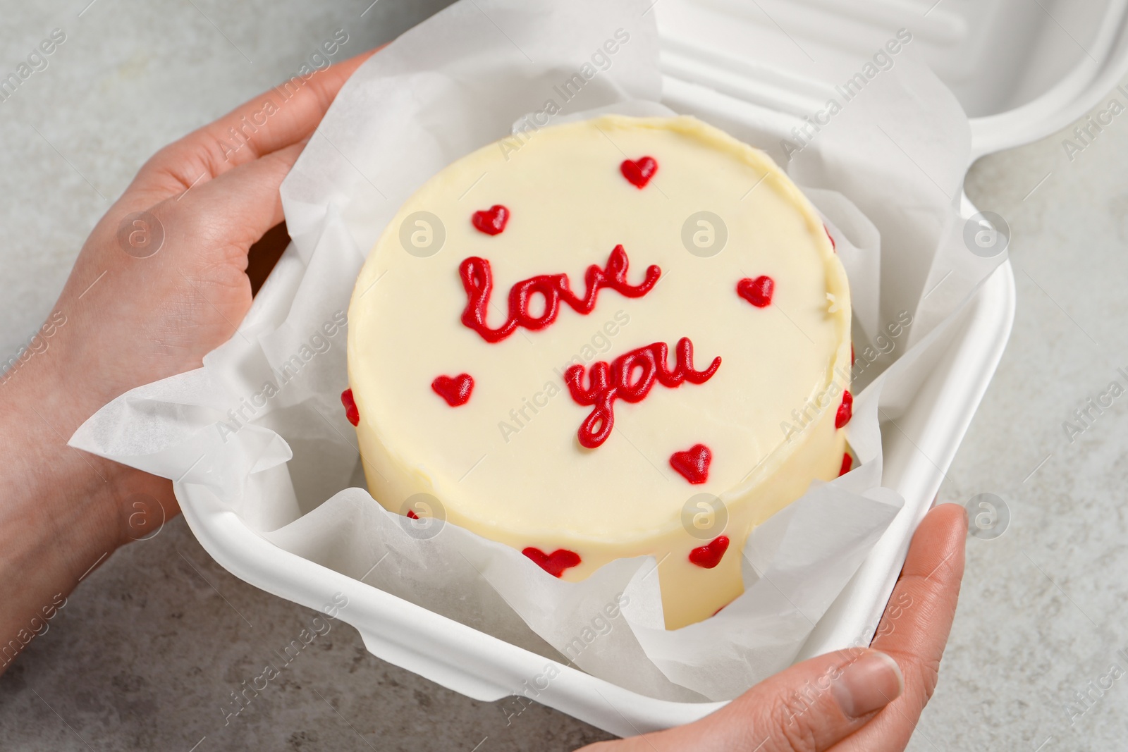 Photo of Woman holding takeaway box with bento cake at light grey table, closeup. St. Valentine's day surprise