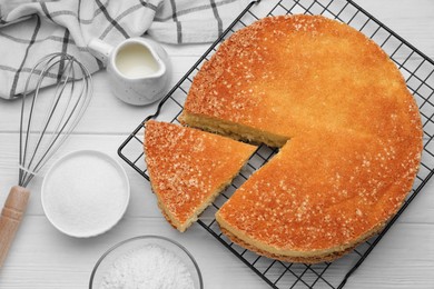 Tasty sponge cake, whisk and ingredients on white wooden table, flat lay