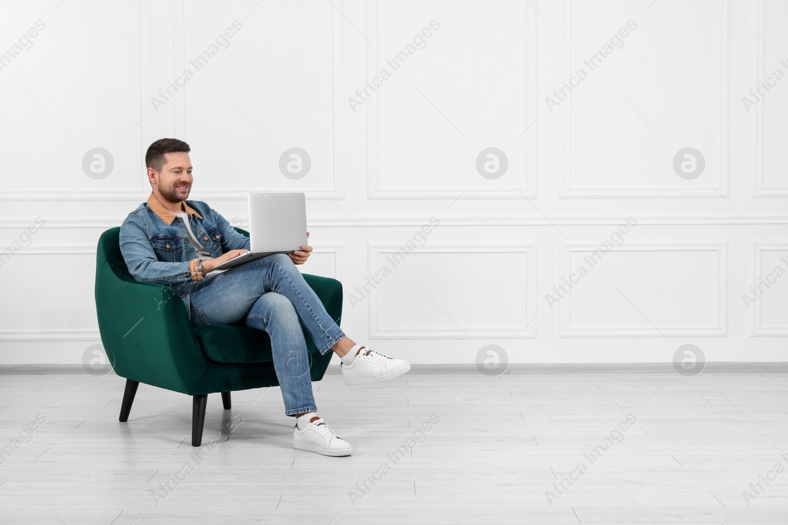 Photo of Happy man sitting in armchair and using laptop indoors, space for text