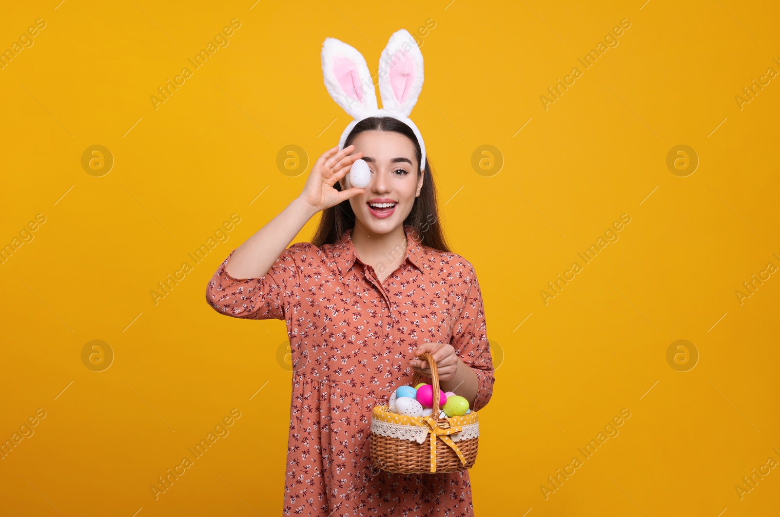 Photo of Happy woman in bunny ears headband holding wicker basket of painted Easter eggs on orange background. Space for text