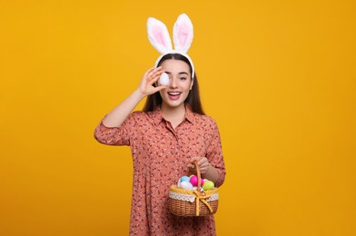 Happy woman in bunny ears headband holding wicker basket of painted Easter eggs on orange background. Space for text