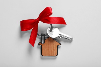 Key with trinket in shape of house and red bow on light grey background, top view. Housewarming party