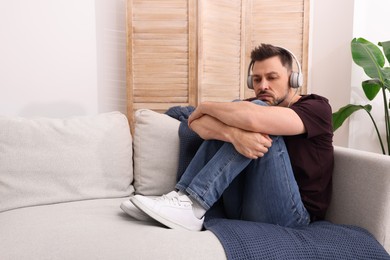 Upset man listening to music through headphones on sofa at home. Loneliness concept