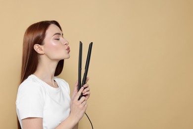 Photo of Beautiful woman with hair iron blowing kiss on beige background, space for text