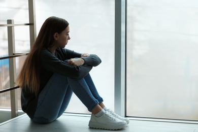 Photo of Upset teenage girl sitting at window indoors. Space for text