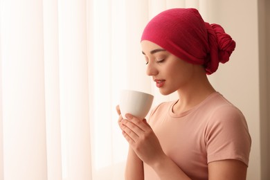 Photo of Cancer patient. Young woman with headscarf and hot drink near window indoors, space for text