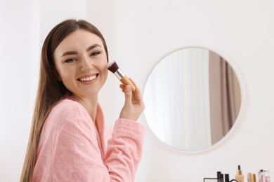Beautiful young woman applying makeup with brush indoors