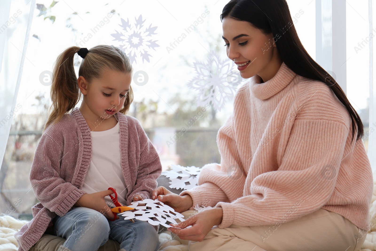 Photo of Mother and daughter making paper snowflake near window at home