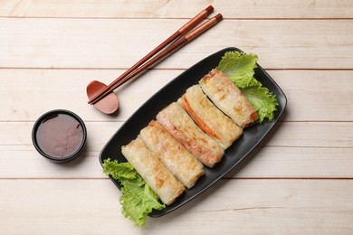 Delicious fried spring rolls, sauce and chopsticks on light wooden table, flat lay