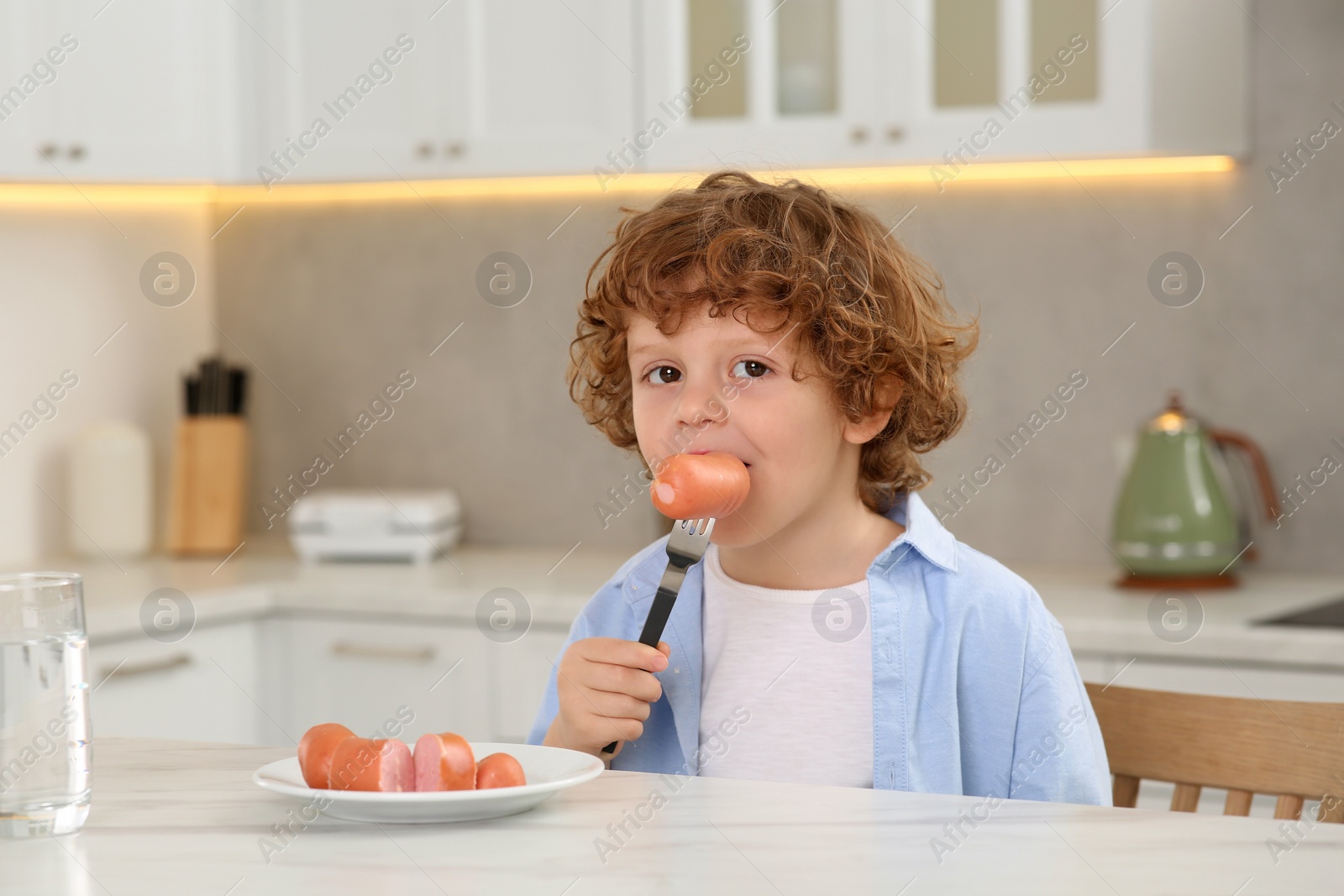 Photo of Cute little boy eating sausages at table in kitchen