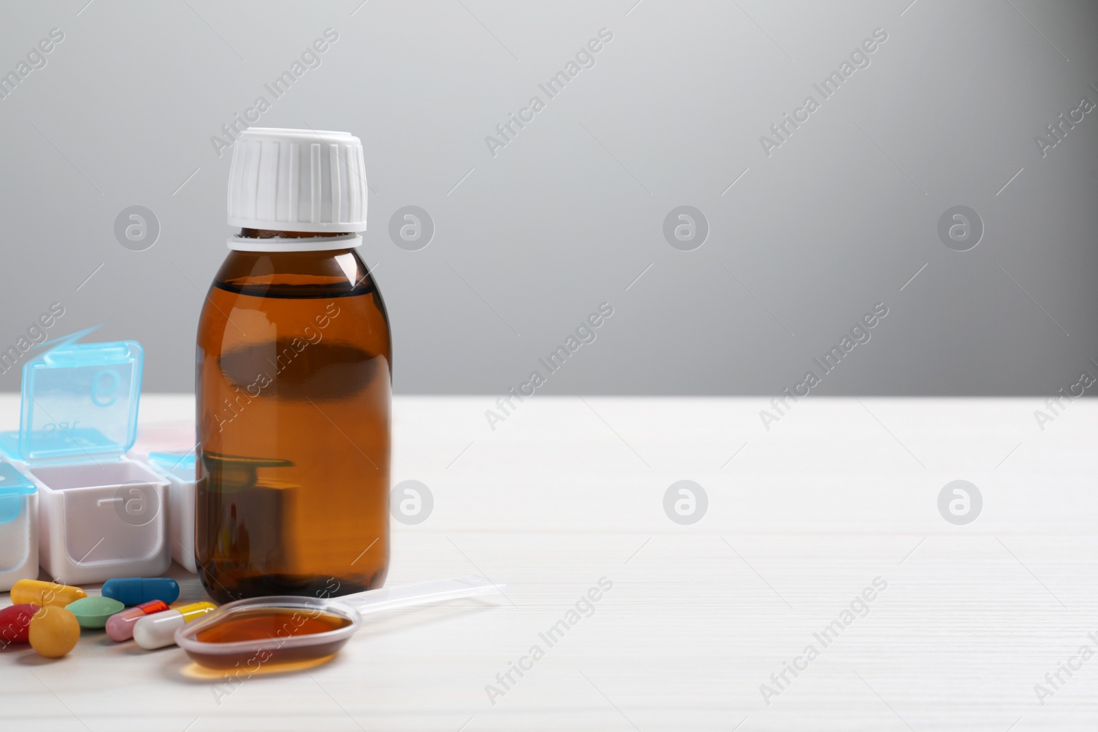 Photo of Bottle of syrup, dosing spoon and pills on white table against light grey background, space for text. Cold medicine