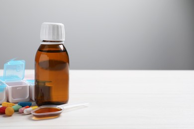 Bottle of syrup, dosing spoon and pills on white table against light grey background, space for text. Cold medicine