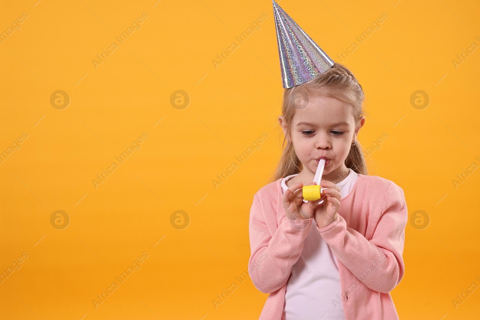 Photo of Birthday celebration. Cute little girl in party hat with blower on orange background, space for text