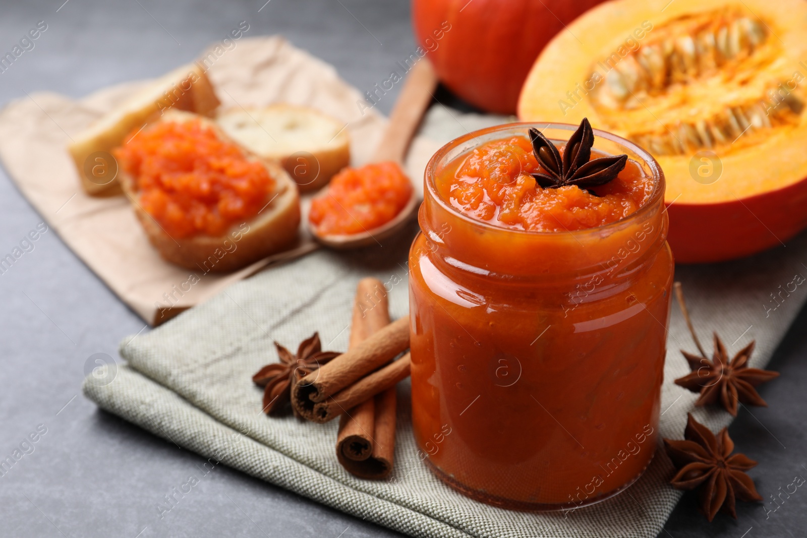 Photo of Jar of pumpkin jam, star anise, cinnamon and fresh pumpkin on grey table, closeup