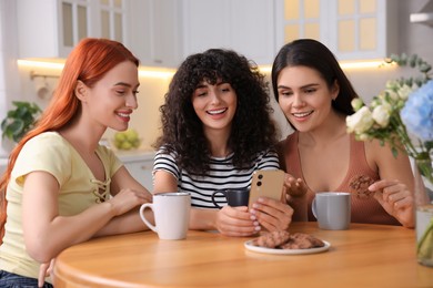 Photo of Happy young friends with smartphone spending time together at table in kitchen