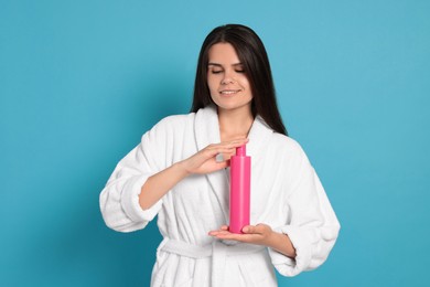 Beautiful young woman in bathrobe holding bottle of shampoo on light blue background