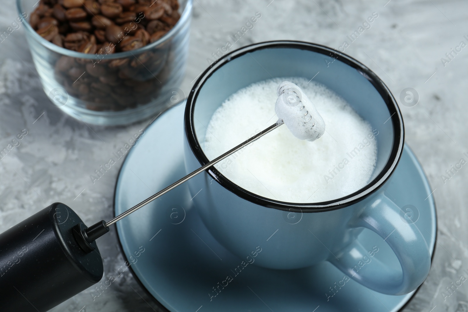 Photo of Mini mixer (milk frother), whipped milk in cup and coffee beans on grey textured table, closeup