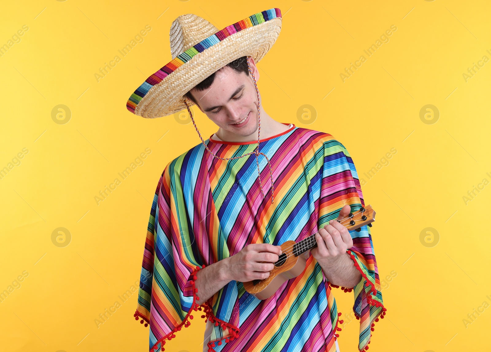 Photo of Young man in Mexican sombrero hat and poncho playing ukulele on yellow background