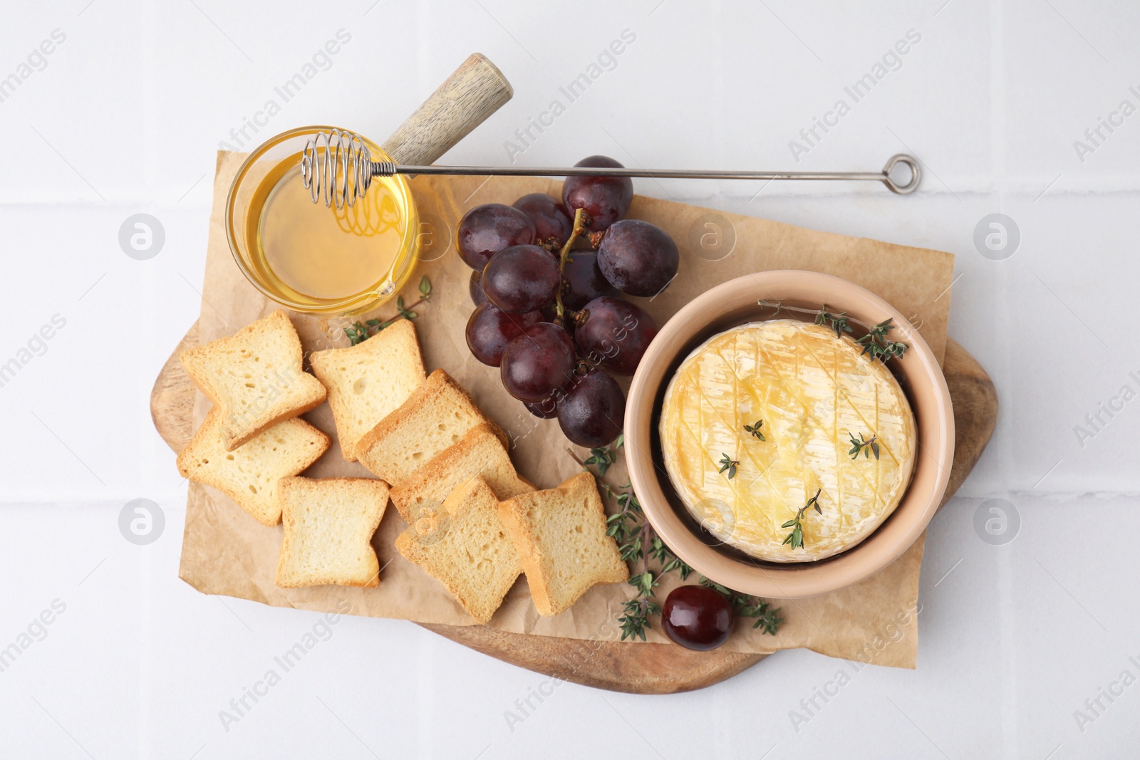 Photo of Tasty baked camembert in bowl, croutons, grapes, honey and thyme on white tiled table, top view