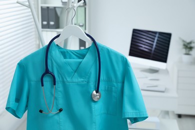 Turquoise medical uniform and stethoscope hanging on rack in clinic, closeup. Space for text