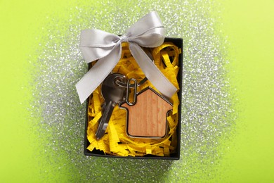 Key with trinket in shape of house, glitter and gift box on light green background, top view. Housewarming party