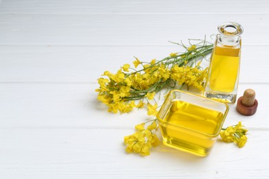 Photo of Rapeseed oil in glass bowl, bottle and beautiful yellow flowers on white wooden table, space for text