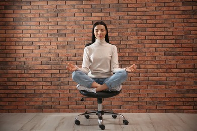 Photo of Young woman meditating on chair near brick wall indoors