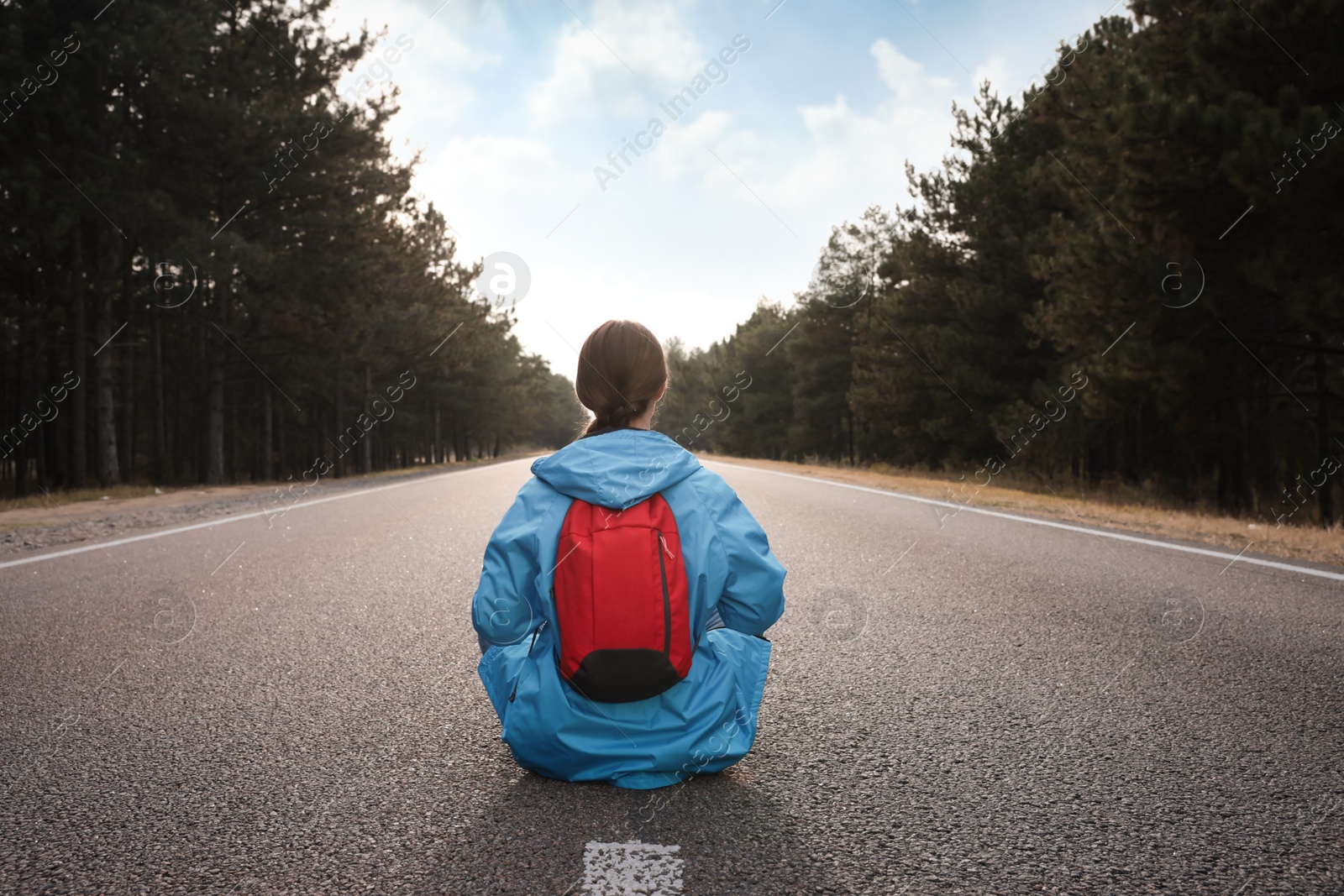 Photo of Young woman with backpack sitting on road near forest, back view