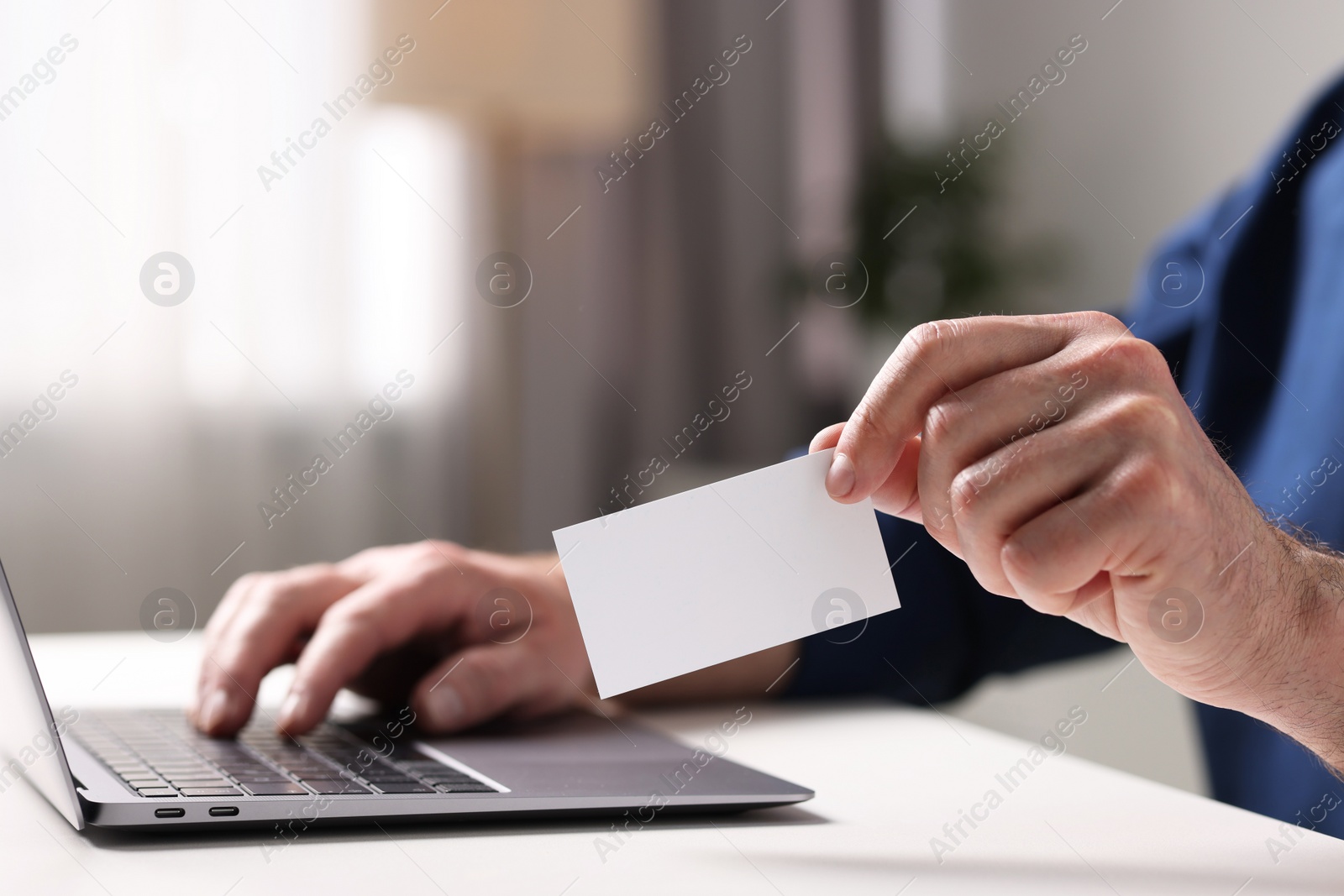 Photo of Man with laptop holding blank business card at white table indoors, closeup