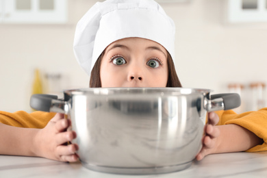 Photo of Surprised little girl wearing chef hat with pot in kitchen