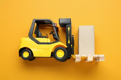 Photo of Toy forklift with wooden pallet and box on orange background, top view
