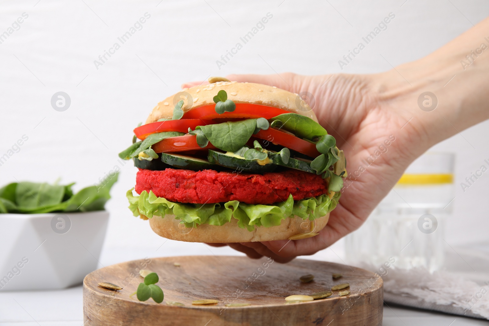 Photo of Woman holding tasty vegan burger with vegetables, patty and microgreens at white table, closeup