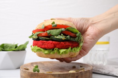 Photo of Woman holding tasty vegan burger with vegetables, patty and microgreens at white table, closeup