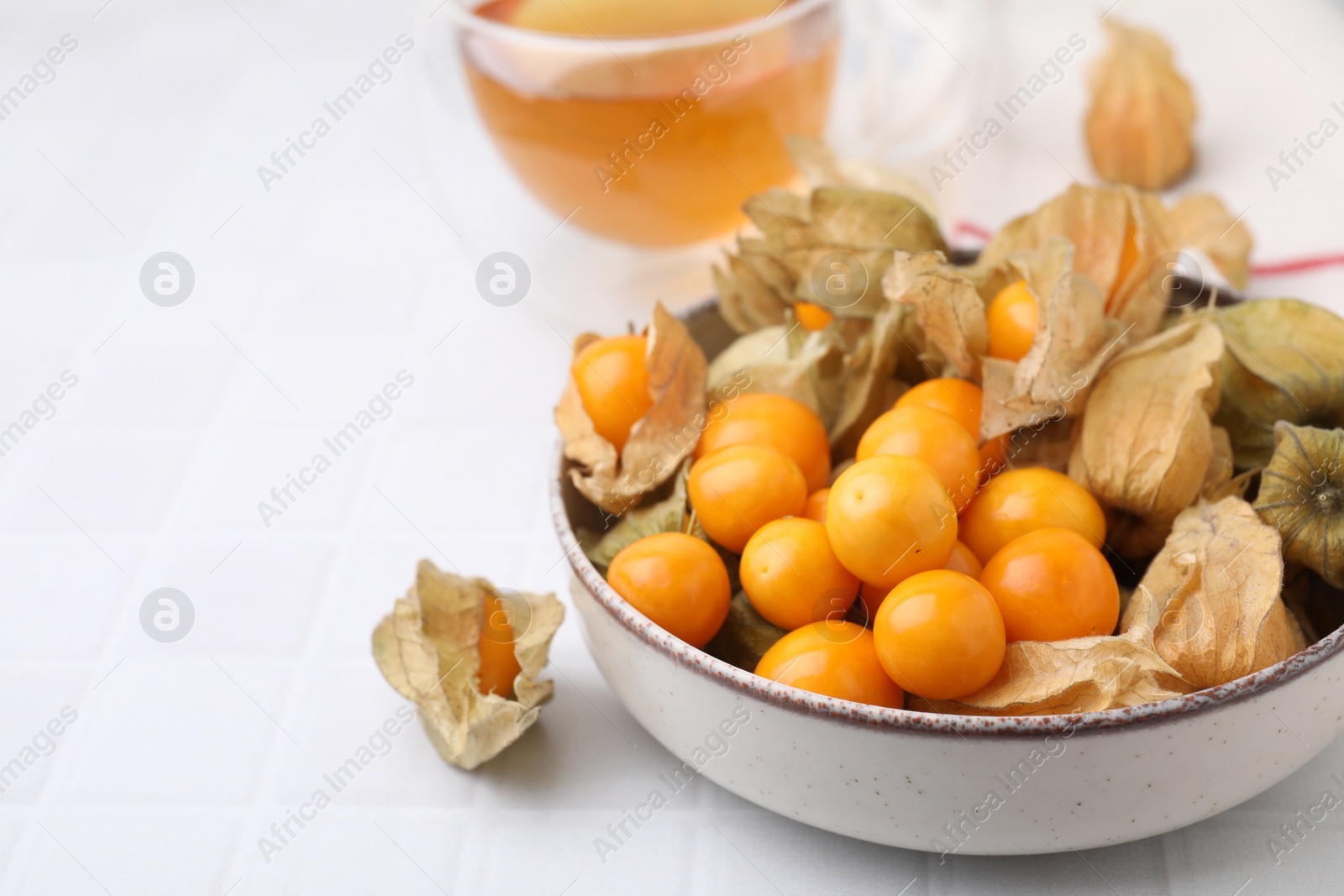 Photo of Ripe physalis fruits with calyxes in bowl on white tiled table, closeup. Space for text