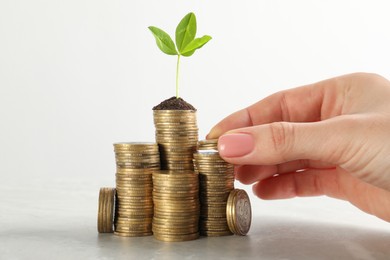 Woman putting coin onto stack with green plant on white marble table, closeup. Investment concept