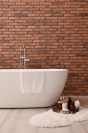 Photo of Modern ceramic bathtub and tray with toiletries near brick wall indoors