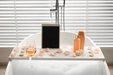 Photo of Wooden tray with tablet, wine, toiletries and flower petals on bathtub in bathroom