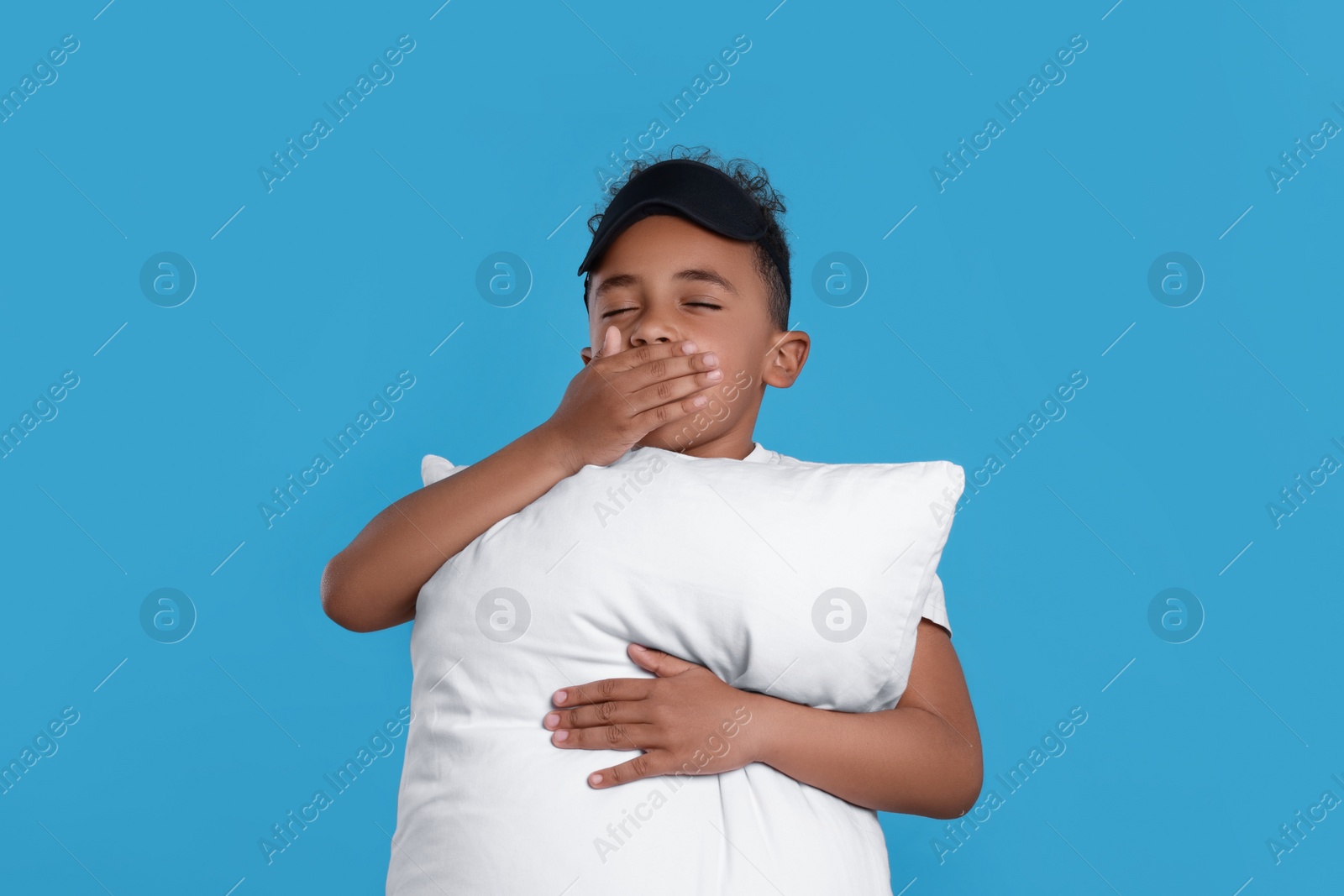 Photo of Boy with pillow and sleep mask yawning on light blue background. Insomnia problem