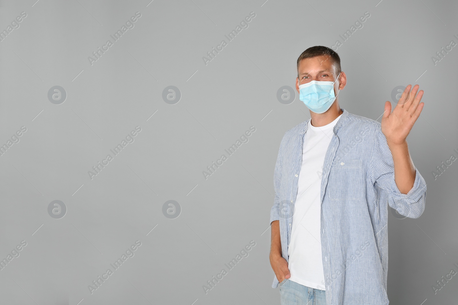 Photo of Man in protective mask showing hello gesture on grey background, space for text. Keeping social distance during coronavirus pandemic