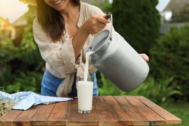 Smiling woman pouring fresh milk from can into glass at wooden table outdoors, closeup