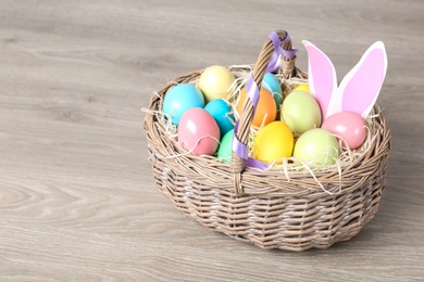 Photo of Wicker basket with Easter eggs and funny bunny ears on wooden table, space for text