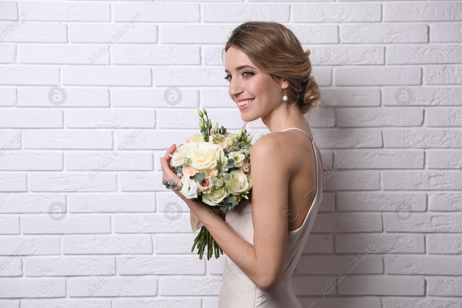 Photo of Young bride with elegant hairstyle holding wedding bouquet near white brick wall