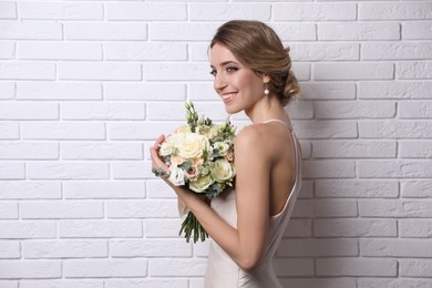 Young bride with elegant hairstyle holding wedding bouquet near white brick wall