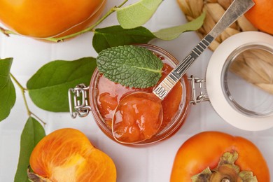 Jar and spoon of tasty persimmon jam, ingredients on white tiled table, flat lay